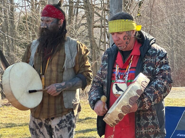 Two men holding hand drums stand next to each other outside. 