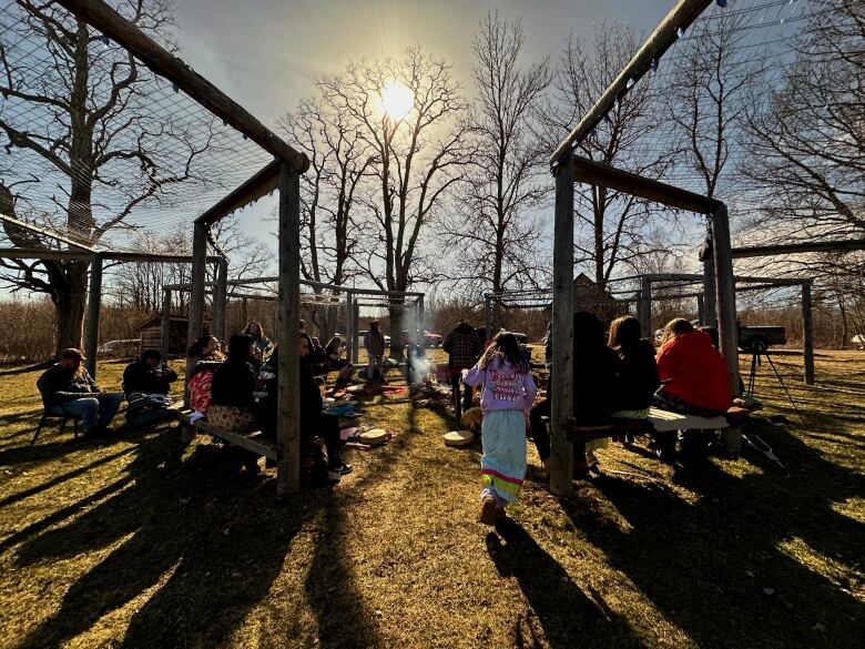 A group of people sit in a circle around a smoking fire pit. The sun casts long shadows across the grass.