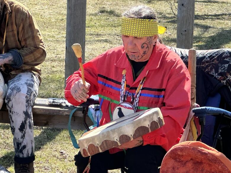 A man wearing a yellow headband and red jacket sits outside on a chair beating a hand drum.