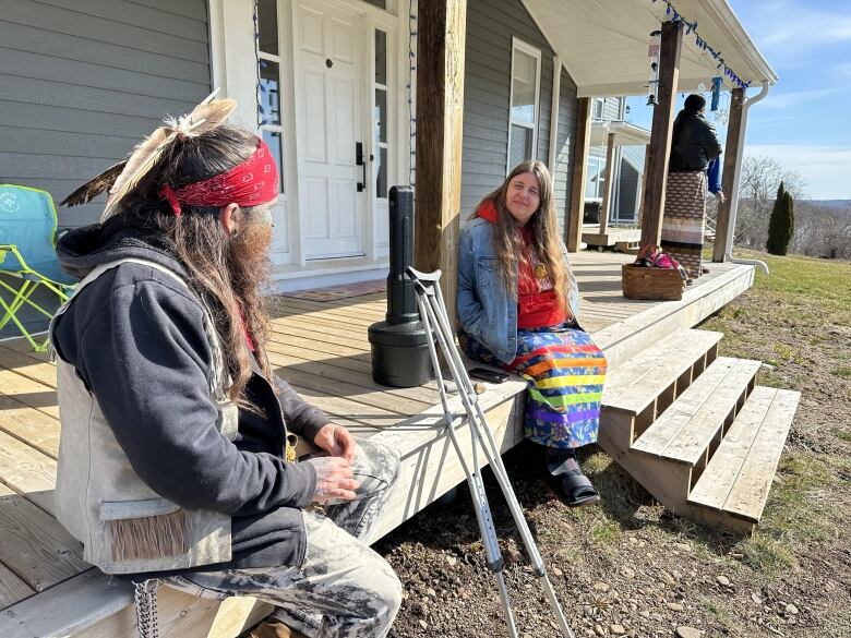 A man wearing a red bandana and a feather in his long, dark hair sits on the front porch of a house in conversation with a woman with long hair and wearing a jean jacket and skirt.