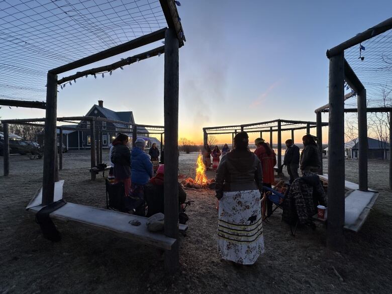 A group of people stand outside in a circle around a fire as the sky darkens above them.