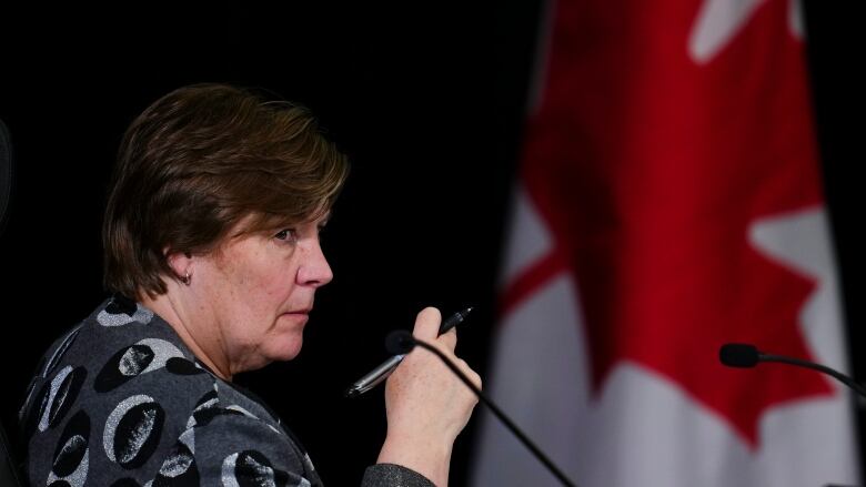 A woman sits in a chair holding a pen. A Canadian flag is drapped in the background.