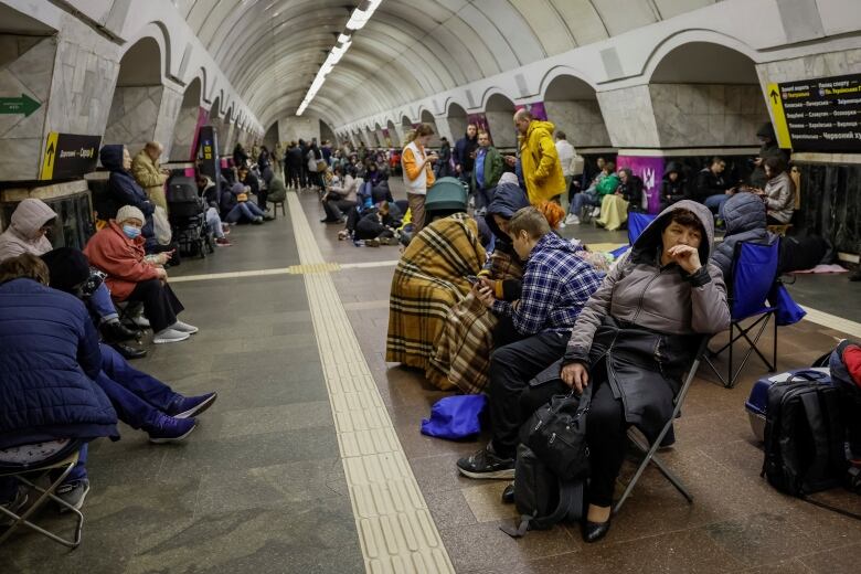 Dozens of people are seen in outerwear, sitting on chairs or on the ground inside a structure that looks like a transit station.