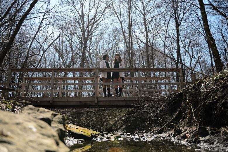 Two girls in white blazers stand on a wooden bridge over a creek.