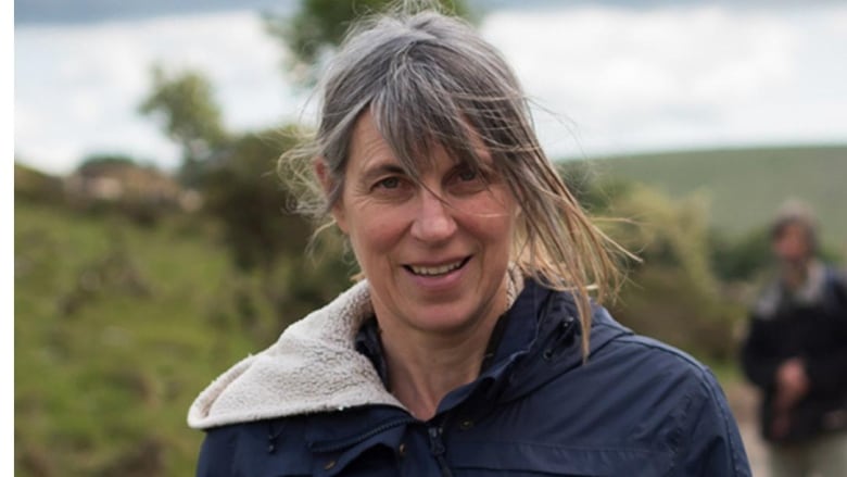 A woman with greying hair in a ponytail in an outdoor landscape. 