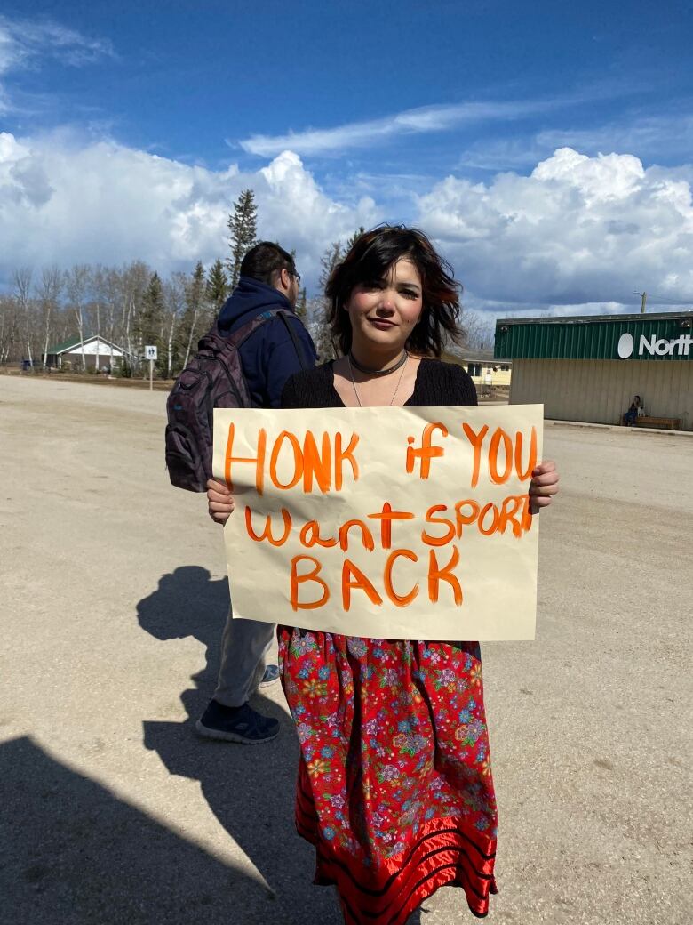 Teen girl in ribbon skirt holds a sign that says 'honk if you want sport back.'