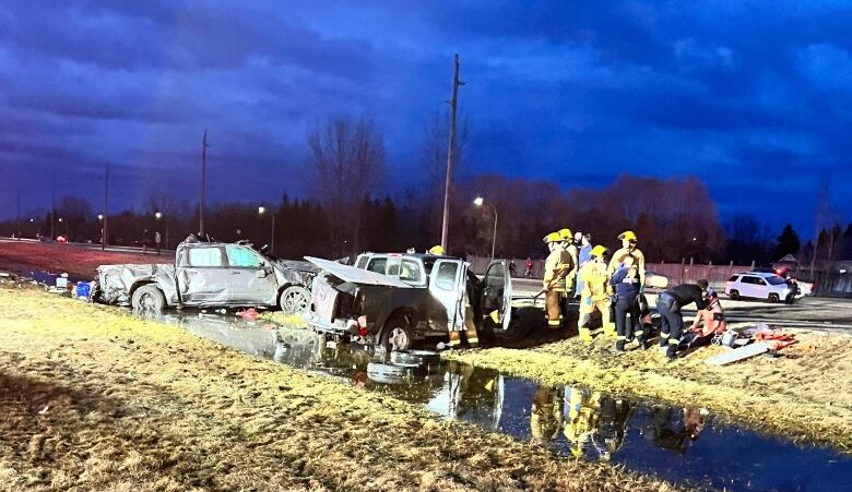 Two bashed-up pickup trucks sit in a country ditch with a little water in it. Emergency workers stand nearby, with some leaning over patients.