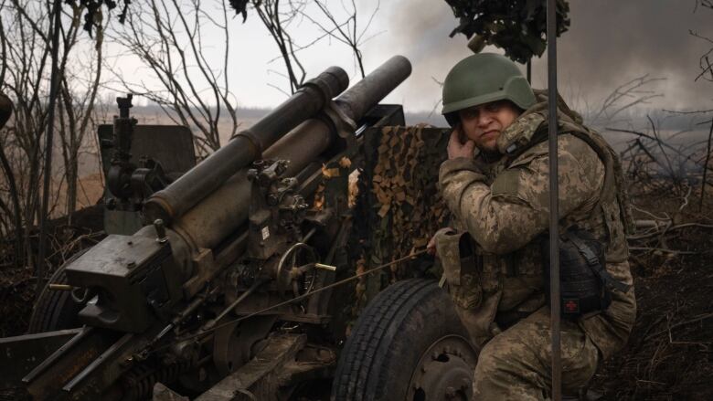 A helmeted soldier wearing head-to-toe camo sits next to a large, wheeled artillery weapon, holding one hand to his ear as smoke billows out from the barrel behind him. 