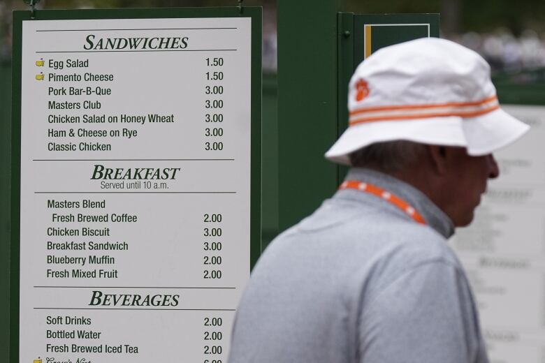 A man wearing a hat walks past a sign listing the prices of food items.