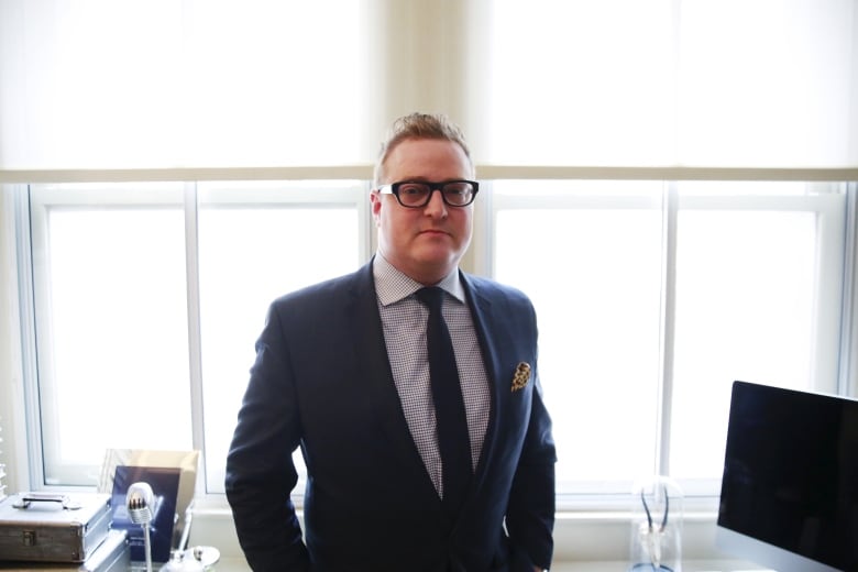 A bespectacled clean-shaven man wearing a suit and tie poses with hands in his pockets in an office setting.