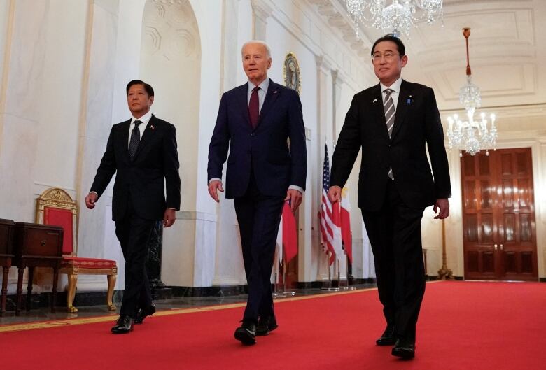 Three men walking on red carpet in white hallway