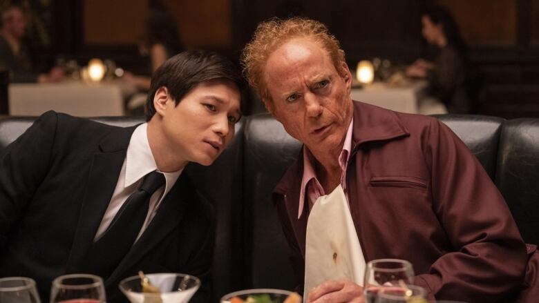 A man wearing a tux leans over to speak with an older man. They are both seated at a dinner table, and are looking at something off frame