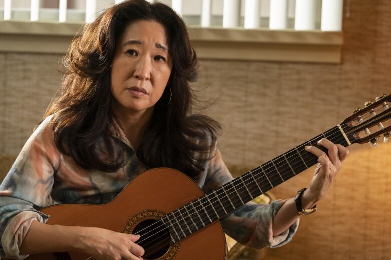 A woman holding a classical guitar looks on. 