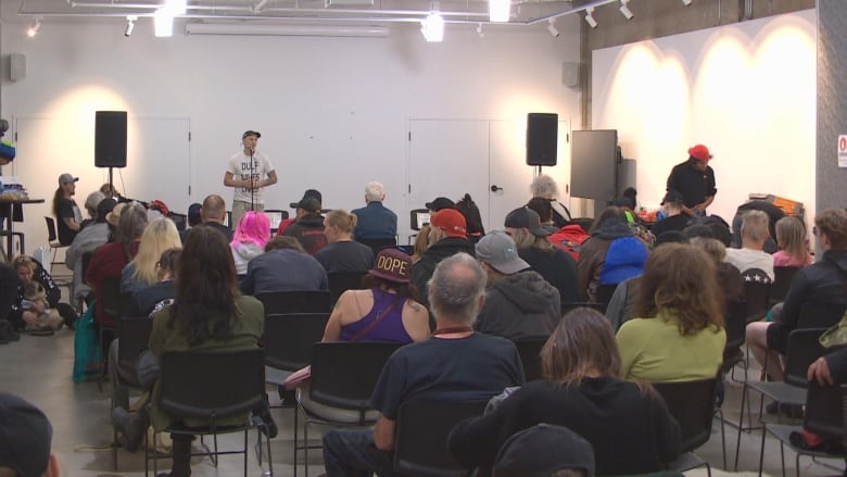 A group of people sit in chairs in a large room facing someone speaking at a microphone.