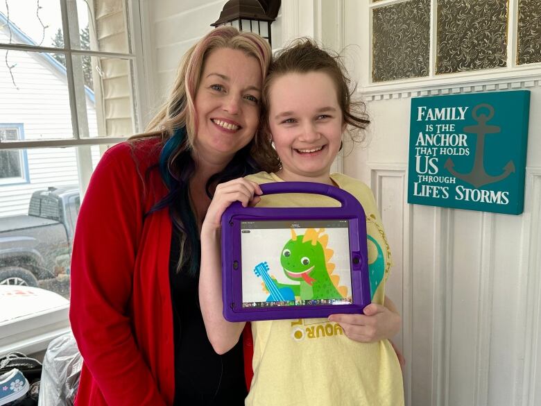 A mother poses with her daughter. The mother wears a bright red shirt. She has blonde hair with blue and pink streaks. Her daughter is holding an iPad with a dragon cartoon. 