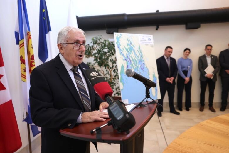 Man wears a suit and glasses. He stands at a podium with flags behind him.