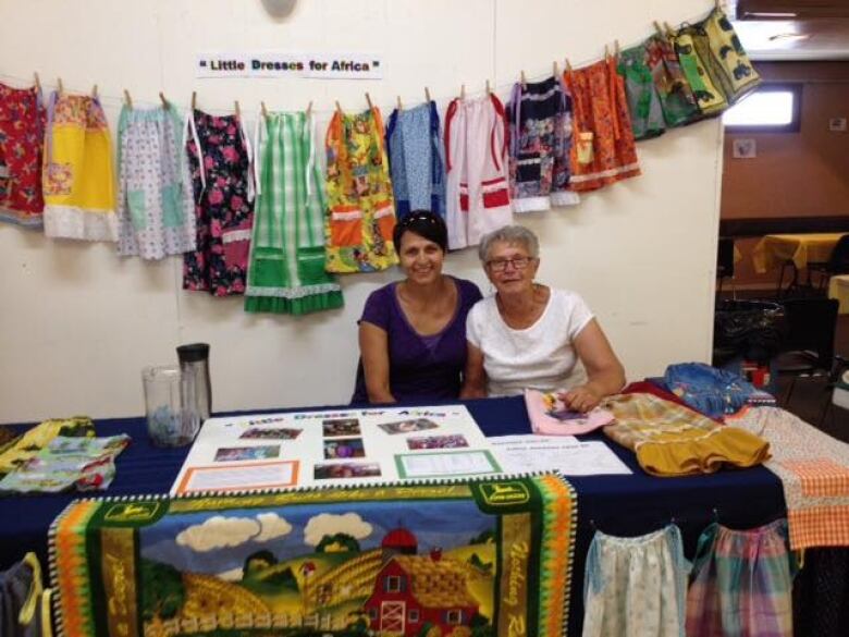 Two women sit at a table with dresses and a poster on it that says 