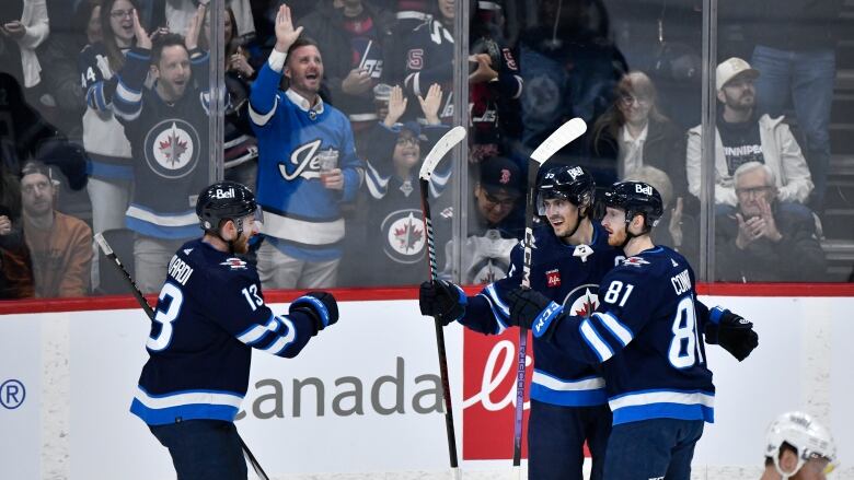 Three Winnipeg Jets players hug in celebration as Jets fans stand up and cheer in the background.