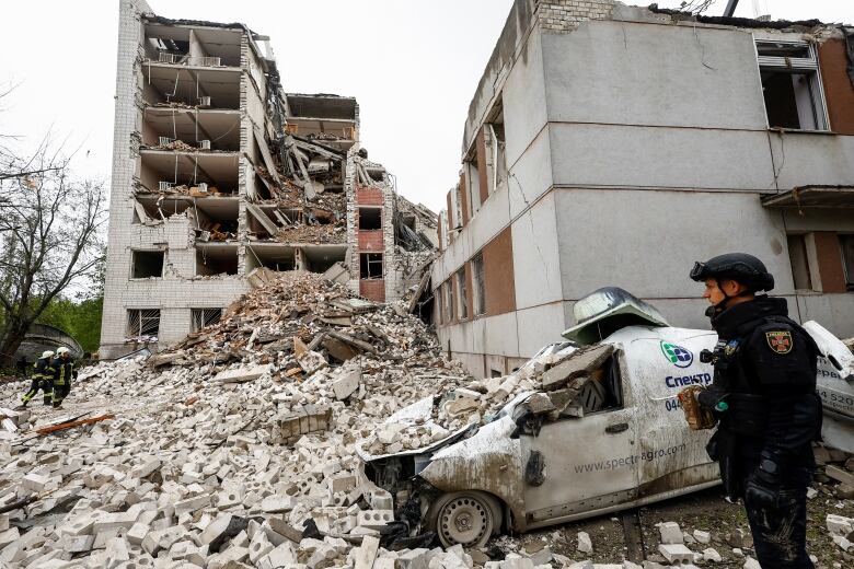 A person wearing a police-style uniform and helmet is shown standing near large piles of concrete debris as a heavily damaged, multi-storey building is shown.