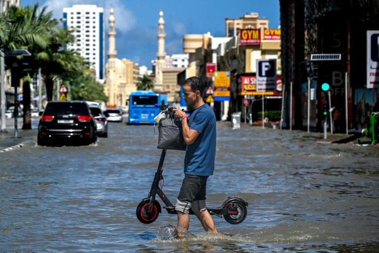 A man in a blue t-shirt and grey shorts carries a scooter across a flooded street.