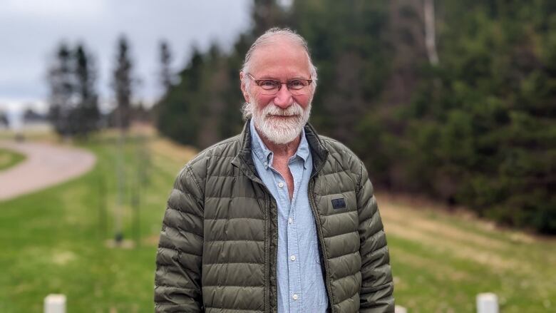 Man with grey beard standing beside trees.