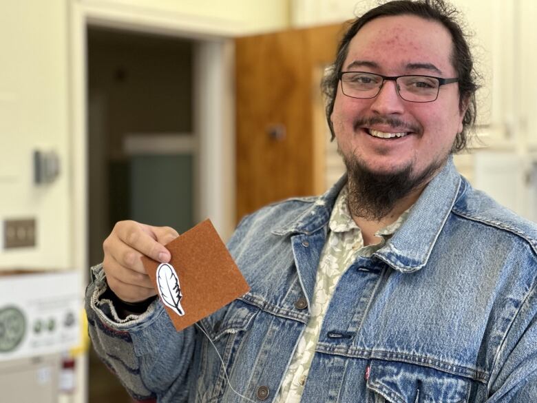 A young, bespectacled person wearing a jean jacket over a floral-print green shirt smiles and holds up a brown square of cloth with a white feather on it.