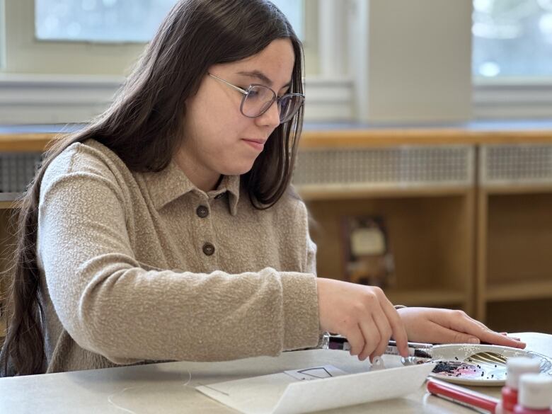 A young, bespectacled person with long dark hair and wearing a beige sweater crafts at a table.