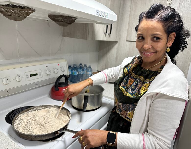 A woman stands next to a stove and stirs flour in a pan.