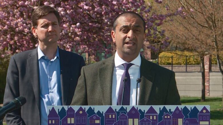 A man stands at a podium and speaks into a microphone. Another man stands behind him.