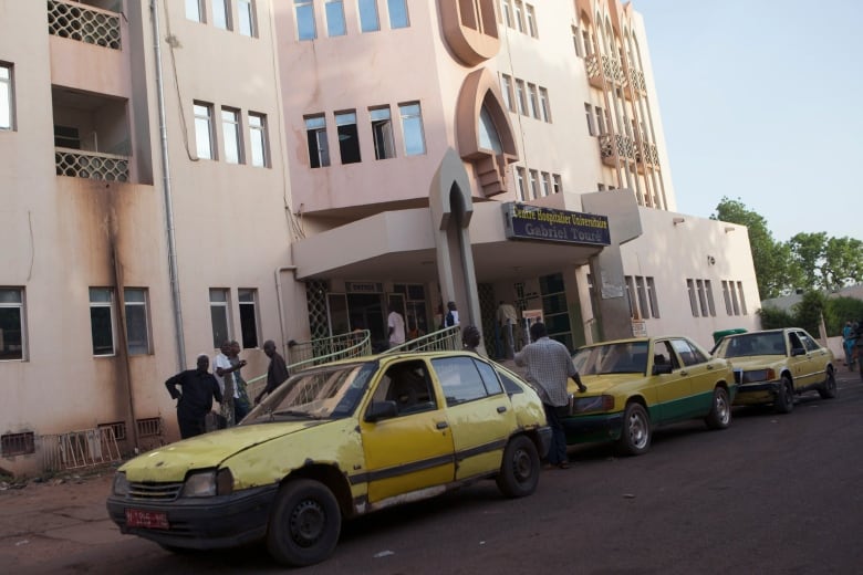 Yellow taxis are parked outside of a large beige building. Several men stand outside.