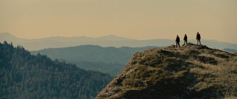 Three silhouetted figures stand on a cliff overlooking a forest.