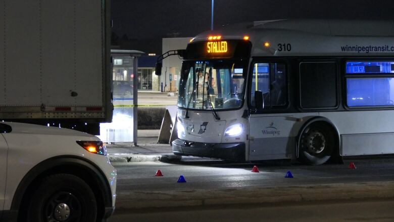 A transit bus is parked at a curb at night time. It's marquee says Stalled. There are police evidence tags on the ground beside it.