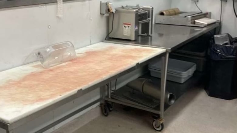 In the foreground is a white table with blood on it, next to a wheeled stainless steel cart with a meat wrapping machine and other equipment. On a lower shelf of the cart are some stacked white plastic bins. There is a garbage bin with a black plastic bag in it to the right of the meat wrapper.