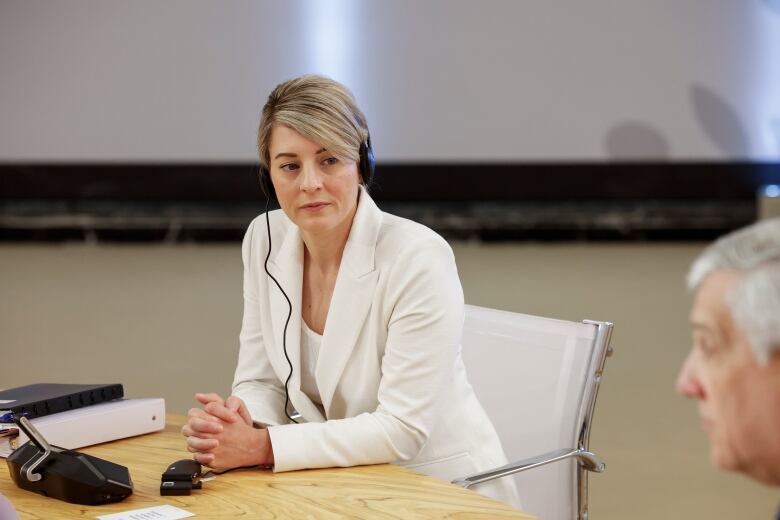 A woman in a white suite sits at a desk with her hands crossed. She listens through an ear piece as a man to her left speaks.