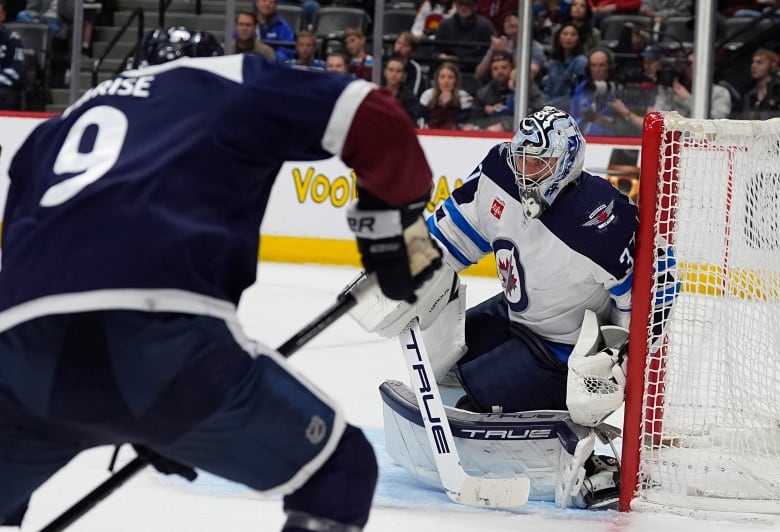 A goalie hugs his left post to face an opposition shooter standing in the foreground.