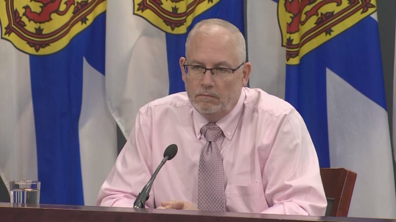 Man in dress shirt sits in front of Nova Scotia flags.