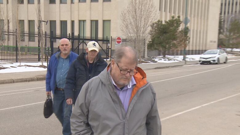 A man in a grey jacket with two men behind him walks away from a courthouse. 