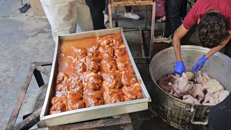 A deep tray on the left with a bunch of chicken and sauce that appear to be uncooked. On the right, a huge pot containing the chicken. A man is leaning in and picking one up.