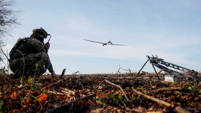 A soldier speaks into a walkie talkie while crouching as a drone flies.