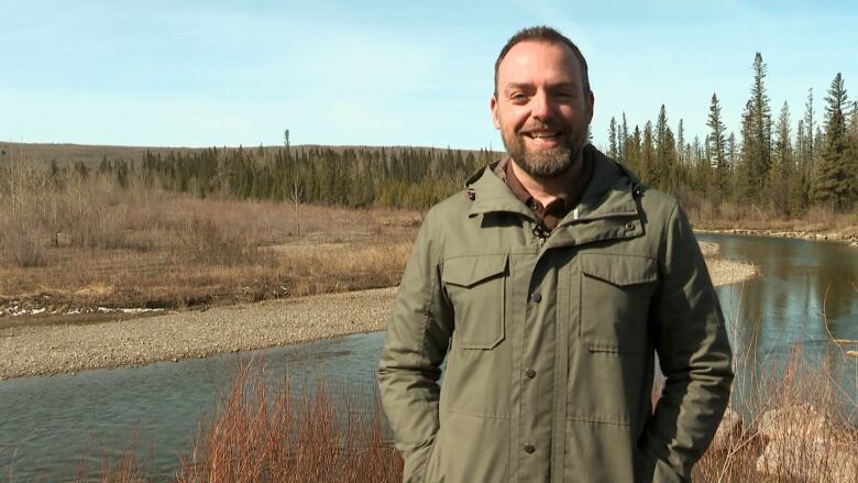 A man wearing a jacket stands in front of a river.