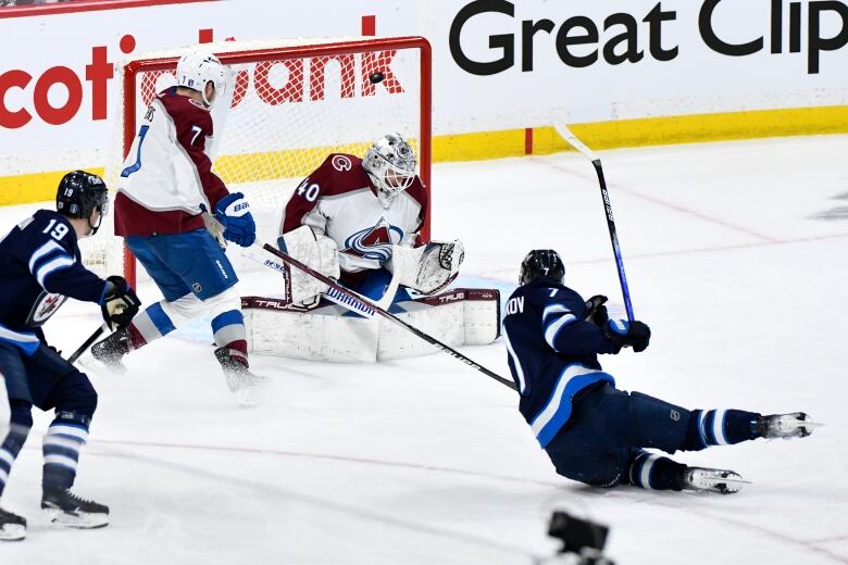 A hockey player makes a swinging motion while falling, as the puck can be seen flying toward the net.