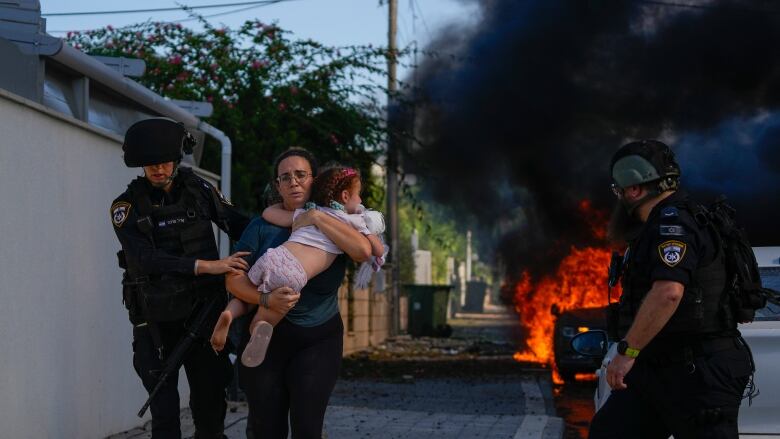 A woman carries a small child in her arms, with police officers in helmets on either side on a sidewalk. Behind them a large ball of fire and smoke is shown in the background.