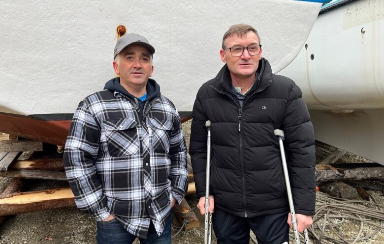 two men, one on crutches, stand in front of a fishing vessel