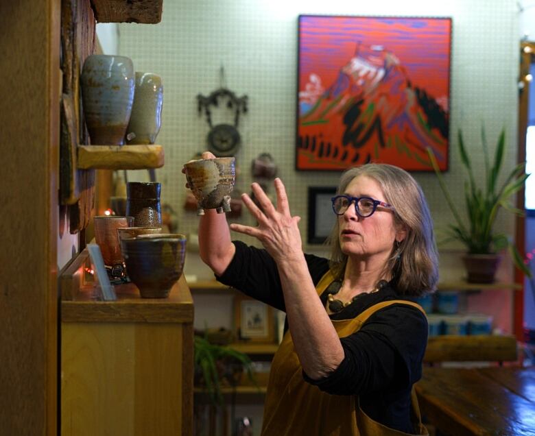 A woman with grey hair wearing a brown apron creates pottery on a pottery wheel.