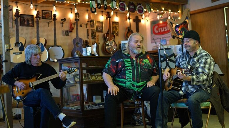 Three men play instruments and sing to six people indoors a space with pottery and musical instruments on the walls and shelves for sale.