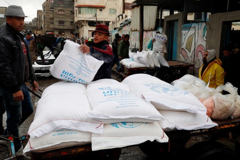 A man piles a white sack of food on top of other sacks loaded onto a trailer. 