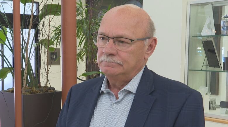 Balding man with glasses and a white mustache in a grey jacket and button-up short stands in a hotel lobby.