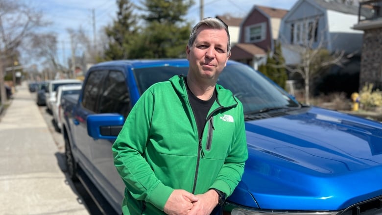 A man in a green jacket stands in front of a blue truck.