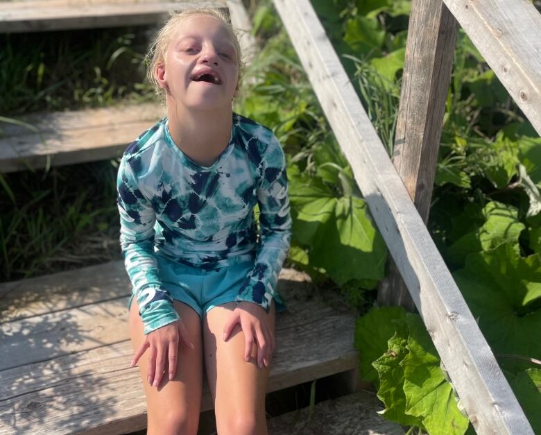 A young girl with blonde hair is smiling on a wooden step. She's wearing a blue outfit. 