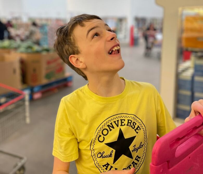 A young boy wears a yellow converse shirt and holds a pink ipad. He is standing in a grocery store. 